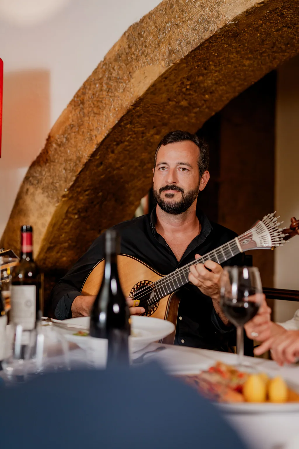 Gitarrist mit der portugiesischen Gitarre unter dem Steinbogen, eine Flasche Wein auf dem Tisch